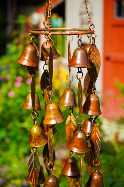 A hanging wind chime made of multiple small metallic bells and leaf-shaped pieces, photographed outdoors against a blurred garden and red-orange building background.