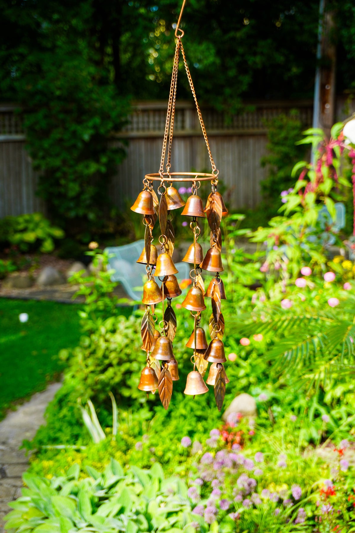 A hanging wind chime with multiple small metallic bells and leaf charms, suspended in a lush green garden with flowers and a fence in the background.