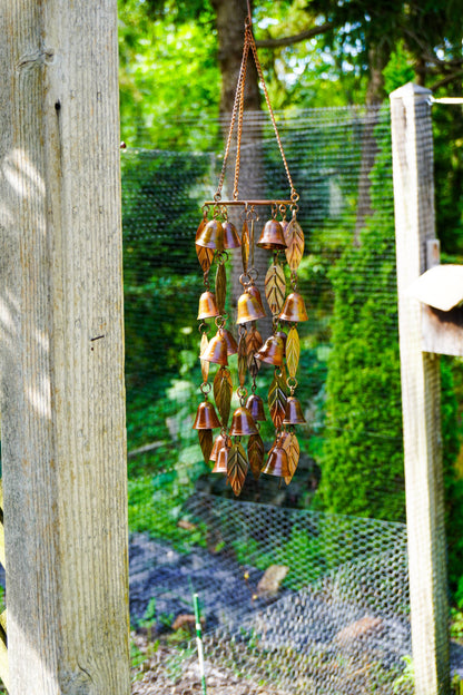 A hanging copper wind chime with small bell-like ornaments and leaf-shaped accents, suspended outdoors between wooden posts with a wire fence and green garden background.