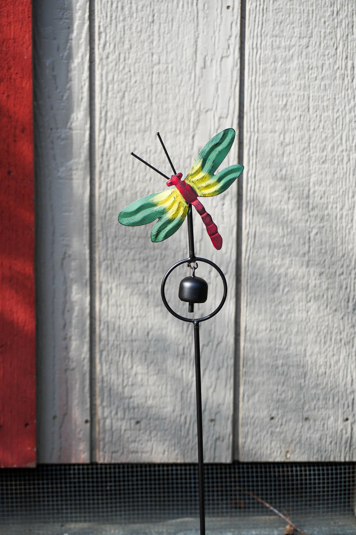 A metal dragonfly garden stake with a small hanging bell, featuring a painted dragonfly with green, yellow, and red wings and body, set against a white wooden wall background.