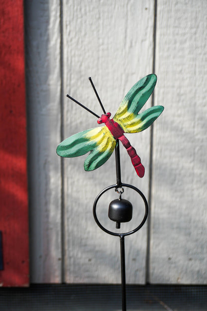 A painted metal dragonfly garden stake with a small hanging bell, shown outdoors against a white wood plank wall and a red vertical panel.