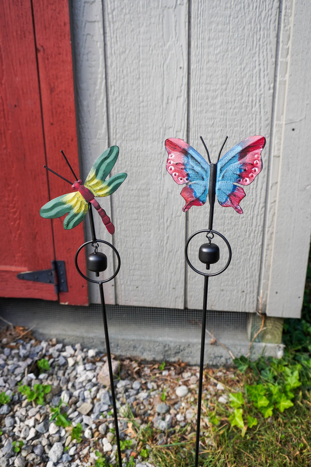 Two decorative metal garden stakes with hanging bells, one shaped like a green and yellow dragonfly and the other like a blue, pink, and red butterfly, placed outdoors in front of a gray wood wall and red door.