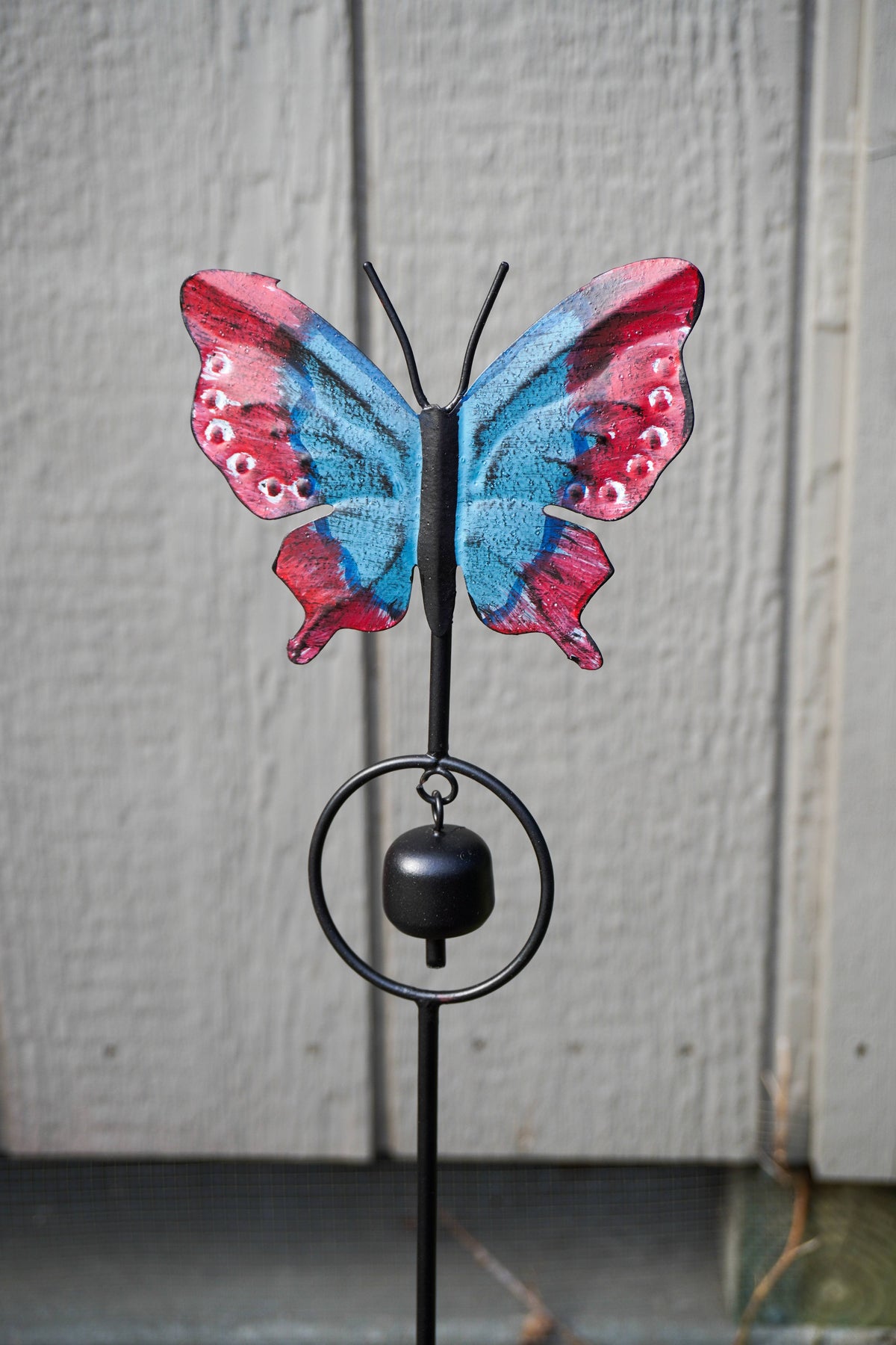A butterfly-shaped metal garden stake with a hanging bell, featuring red and blue patterned wings, photographed against a gray wooden fence.
