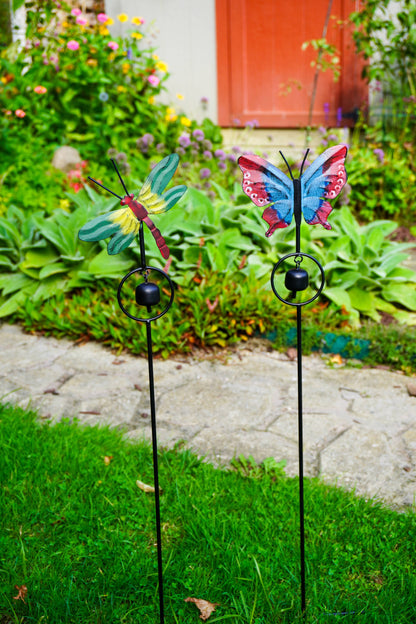 Two metal garden stakes with colorful winged ornaments, one dragonfly and one butterfly, displayed in a green garden next to a stone path.