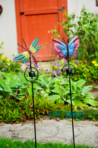 Two colorful metal garden stake wind chimes shaped like a green and yellow dragonfly and a pink, blue, and purple butterfly, set in a garden with a red door in the background.