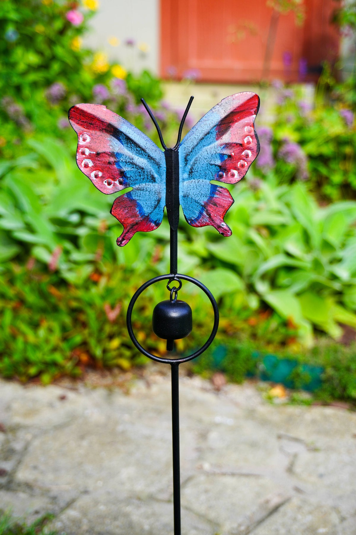A butterfly-shaped garden stake wind chime with a black metal stake, circular ring, and hanging bell, topped with a colorful butterfly wing design in blue, pink, and red, set against a blurred garden background.