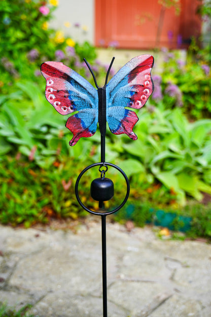 A butterfly-shaped garden stake wind chime with a black metal stake, circular ring, and hanging bell, topped with a colorful butterfly wing design in blue, pink, and red, set against a blurred garden background.