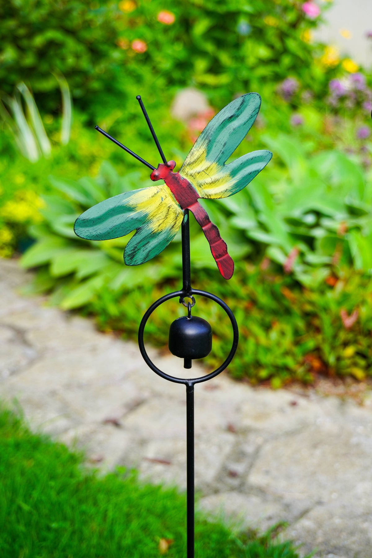 A metal dragonfly garden stake with a small hanging bell, photographed outdoors in a garden. The dragonfly has green wings, a red and yellow body, and a black stake and ring stand against a blurred green garden background.