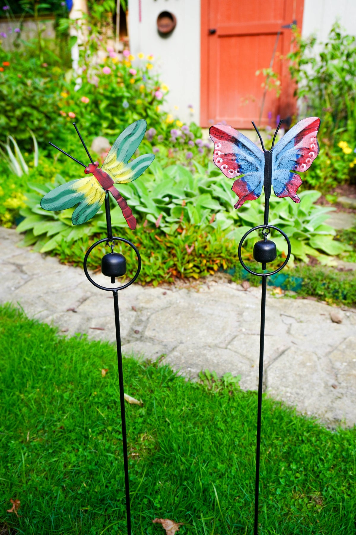 Two colorful metal garden stakes with butterfly and dragonfly designs, placed in a green lawn in front of a garden bed and stone path.