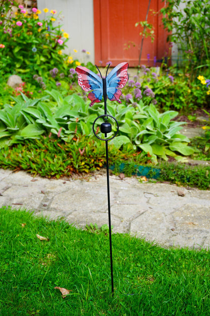 A butterfly-shaped garden stake with a blue, red, and white wing design stands in a green lawn beside a stone path and flower beds.