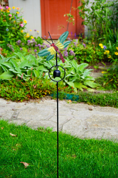 A garden stake featuring a metal dragonfly ornament with green wings, a red body, and a small black bell, set in a grassy garden with a stone path and plants in the background.
