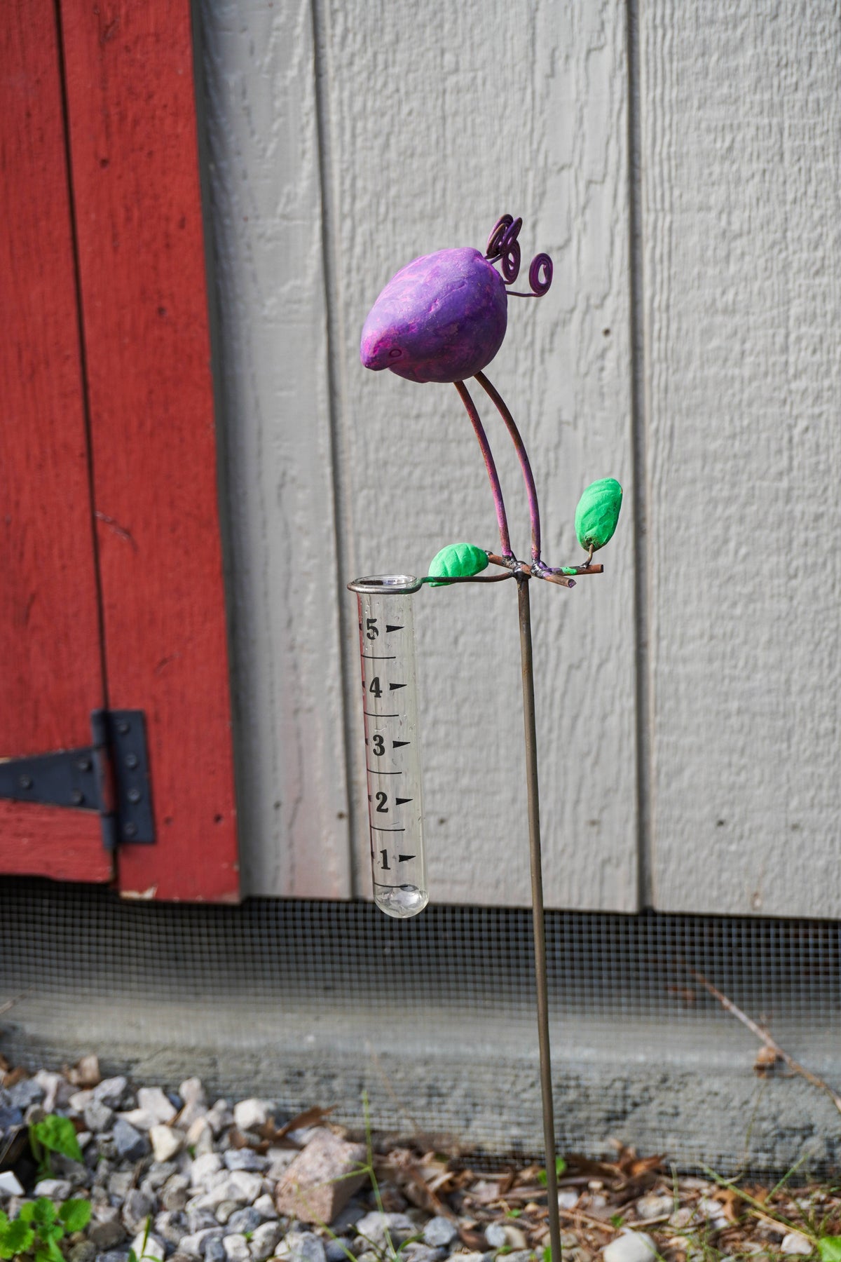 A metal garden stake sculpture shaped like a purple bird or pod with green leaves, mounted on a clear graduated cylinder measuring cup, set outdoors against a gray wood siding wall and red shutters.