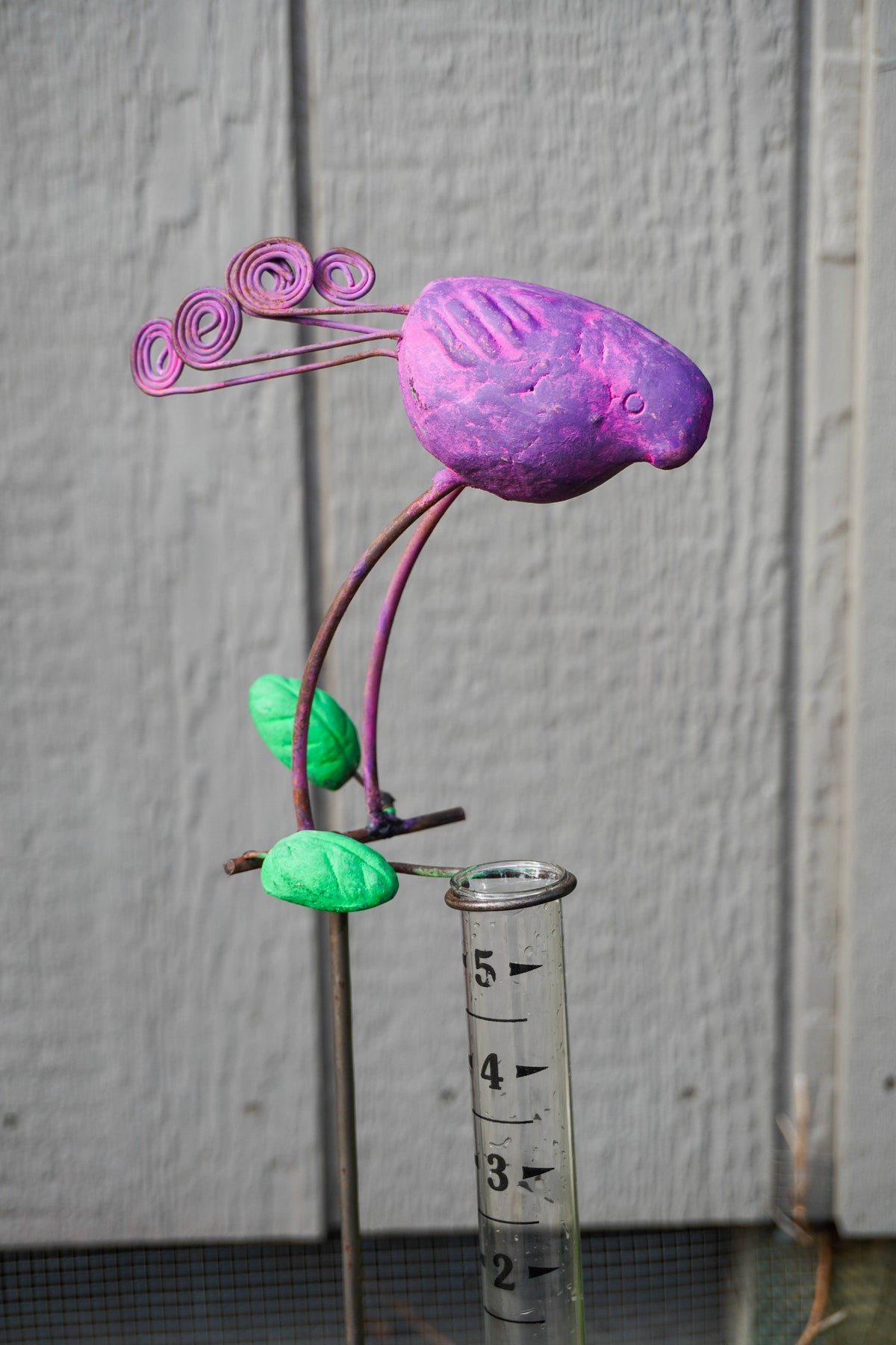 A handmade metal garden sculpture of a flower with a purple bloom, green leaves, and pink curled petals on a metal stake, shown next to a clear plastic measuring cup against a gray wooden background.