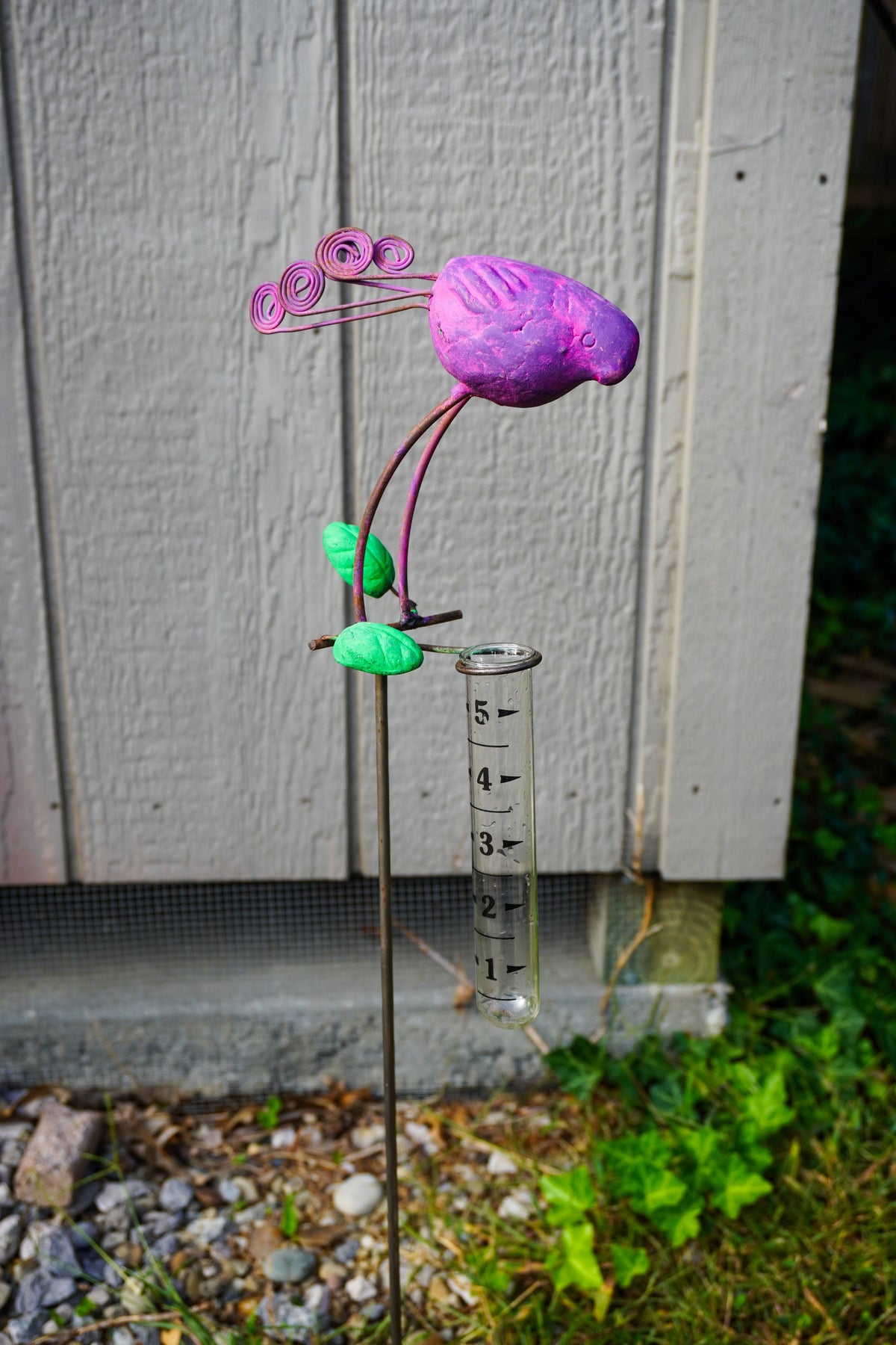A metal flower garden stake sculpture with a purple bird head, green leaves, and a clear graduated cylinder hanging from the stem, photographed outdoors in front of a gray wooden fence.