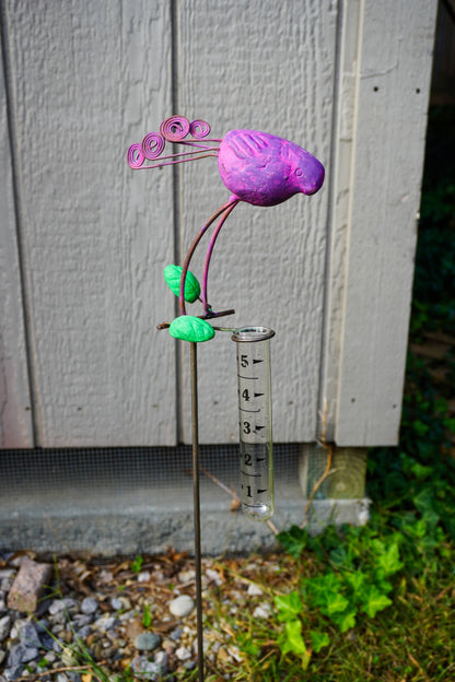 A metal flower garden stake sculpture with a purple bird head, green leaves, and a clear graduated cylinder hanging from the stem, photographed outdoors in front of a gray wooden fence.