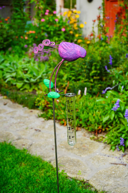 A garden sculpture shaped like a purple bird with green leaves and a hanging clear test tube, photographed outdoors in a garden next to a stone path.