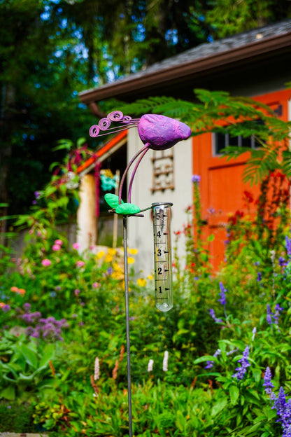 A garden stake sculpture featuring a purple bird head, green stem and leaf details, and a clear graduated tube, set in a lush garden with a red-orange shed in the background.
