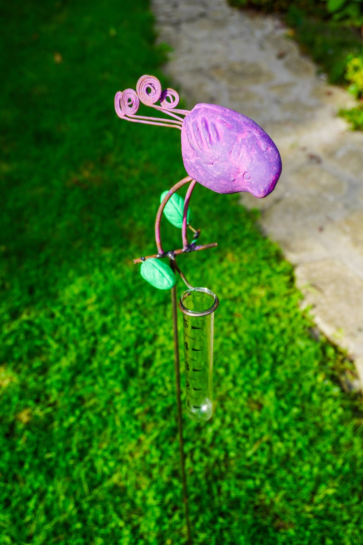 A decorative metal garden stake shaped like a purple bird with green leaves and curled purple stems, mounted on a clear glass test tube, photographed outdoors on grass beside a stone path.