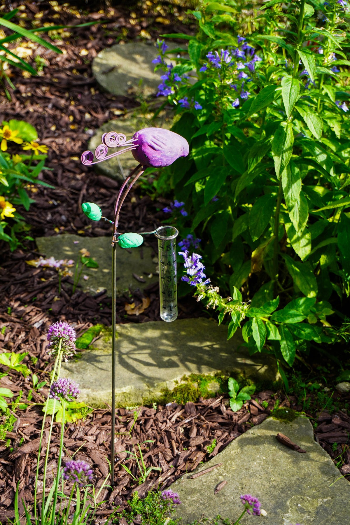 A garden stake sculpture featuring a purple bird head and green leaves, holding a clear glass tube, set in a garden bed with mulch, stones, and green foliage.