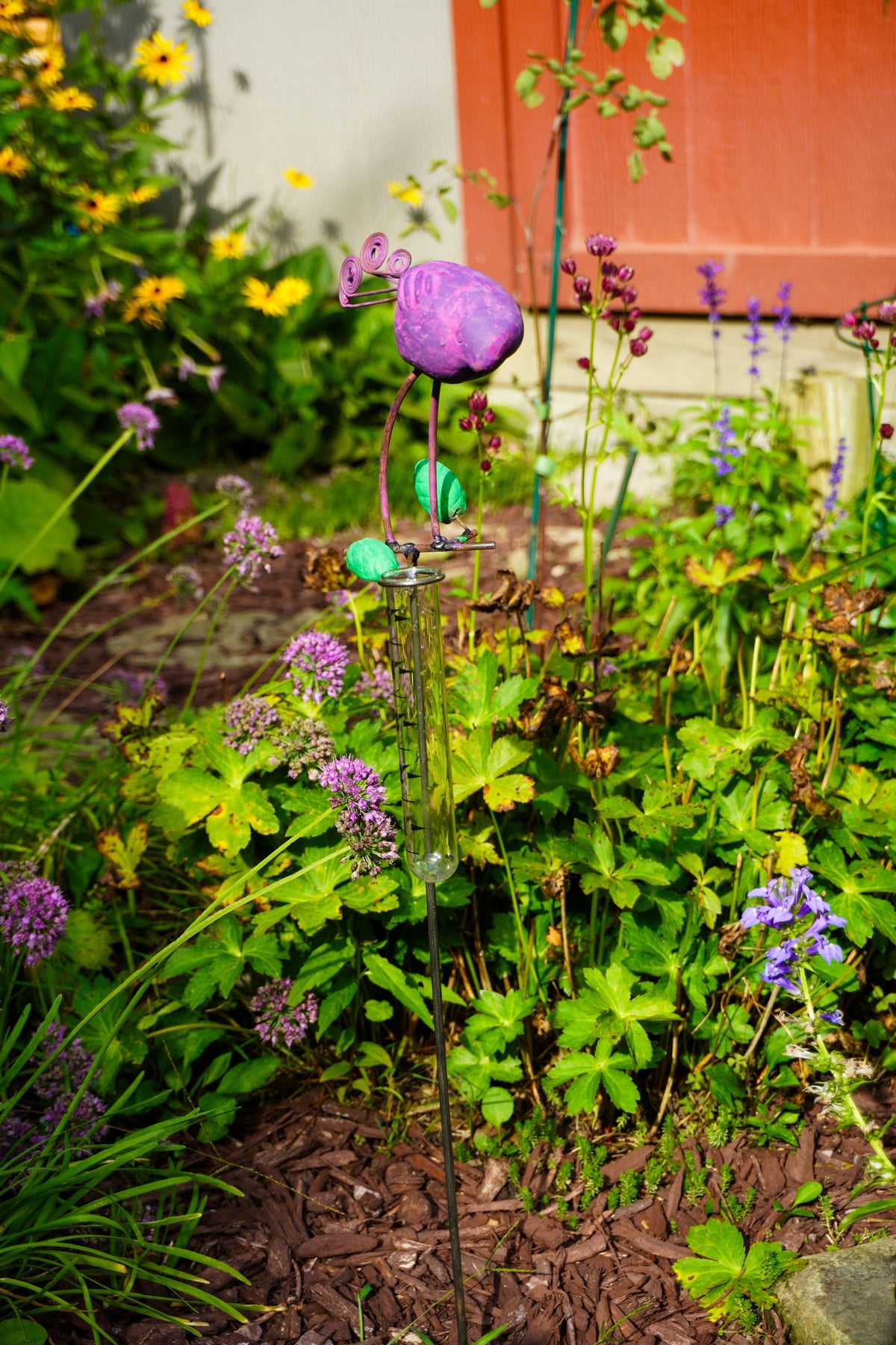 A purple bird-shaped metal garden stake with a butterfly topper and small green accents, planted in a lush garden bed with green foliage and other purple flowers.