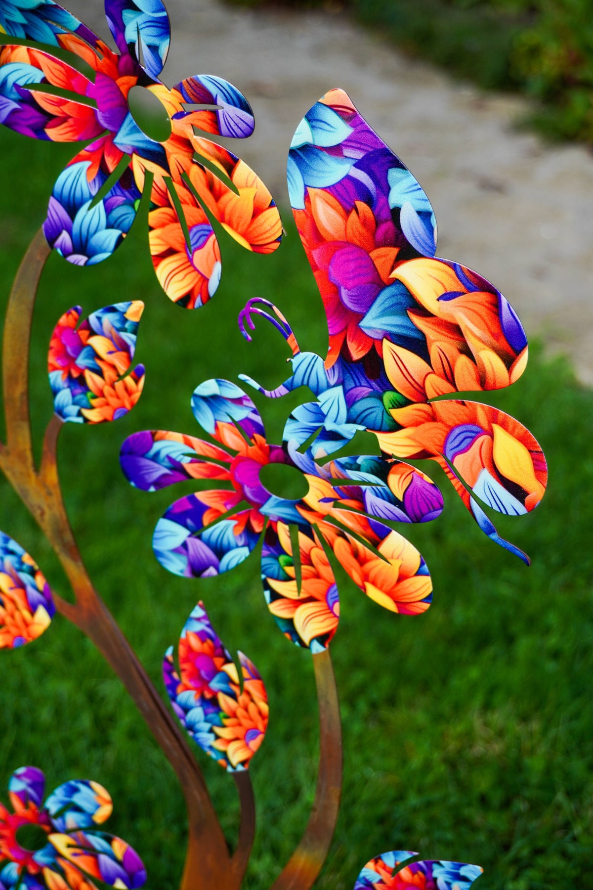Colorful metal garden sculptures shaped like stylized flowers and leaves, featuring vibrant patterns in blue, purple, orange, and yellow, set against a blurred green lawn background.
