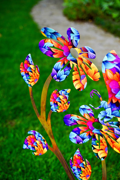Colorful metal garden sculptures shaped like flowers and leaves, featuring a vibrant multicolor pattern of blue, purple, orange, and pink, set against a blurred green lawn and stone path background.