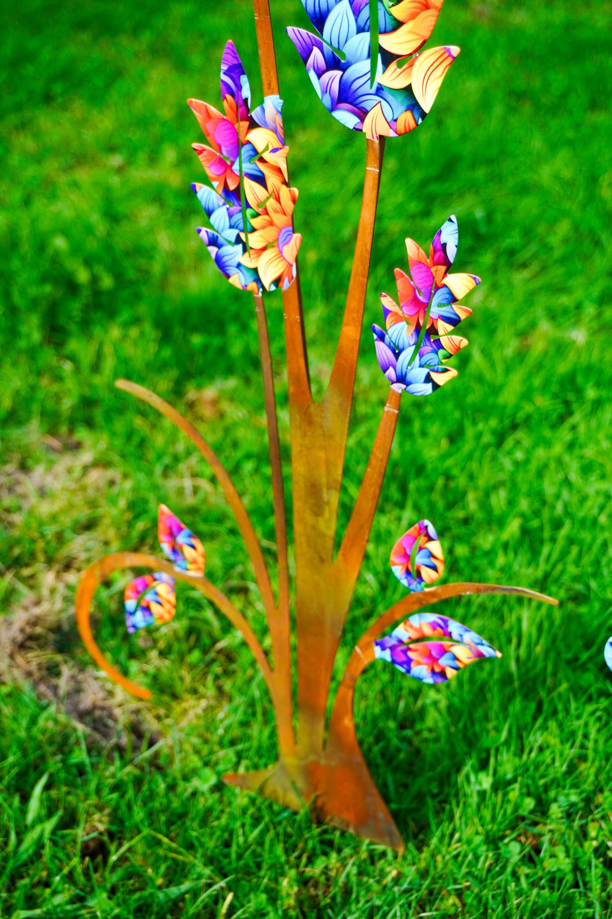 A colorful metal garden stake shaped like stylized flowers with orange-brown stems and bright multicolored petals, photographed outdoors on green grass.
