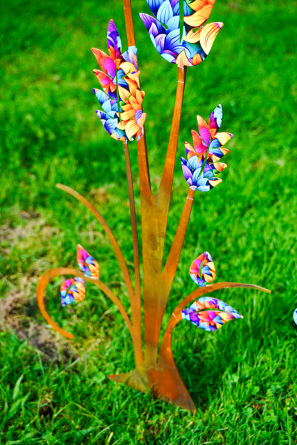 A colorful metal garden stake shaped like stylized flowers with orange-brown stems and bright multicolored petals, photographed outdoors on green grass.
