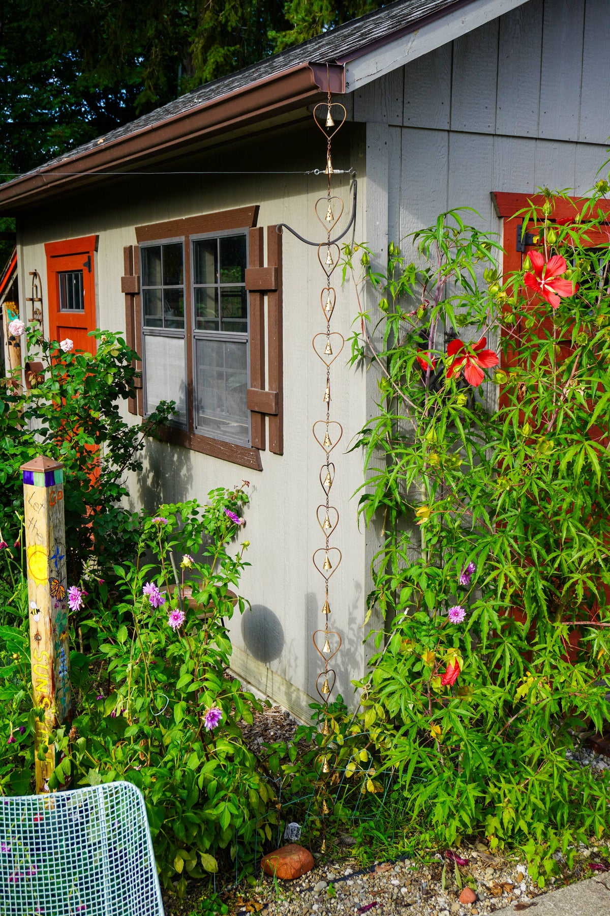 A garden trellis with a heart-shaped metal loop design hanging vertically on the exterior wall of a gray house, surrounded by lush green plants and red flowers.