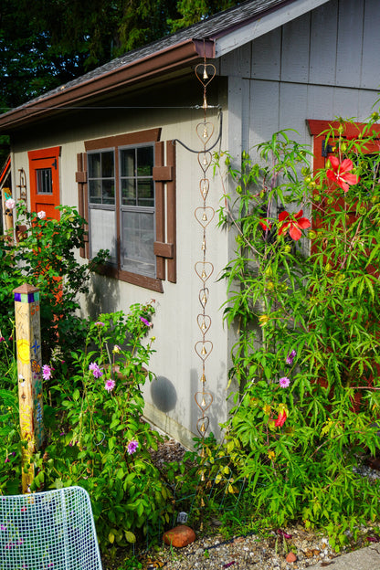 A garden trellis with a heart-shaped metal loop design hanging vertically on the exterior wall of a gray house, surrounded by lush green plants and red flowers.