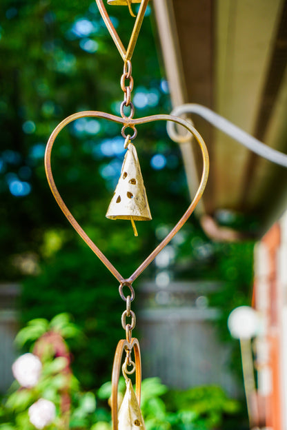 A rustic metal heart-shaped garden wind chime with a small bell in the center, hanging outdoors against a blurred green garden background.
