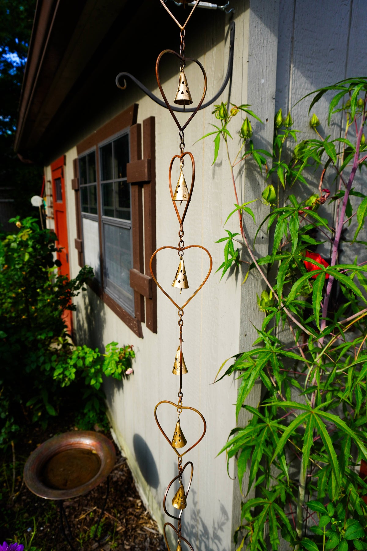 A decorative metal garden wind spinner with a series of heart-shaped wire frames and small hanging bells, shown outdoors against a house wall with green plants and a bird bath nearby.