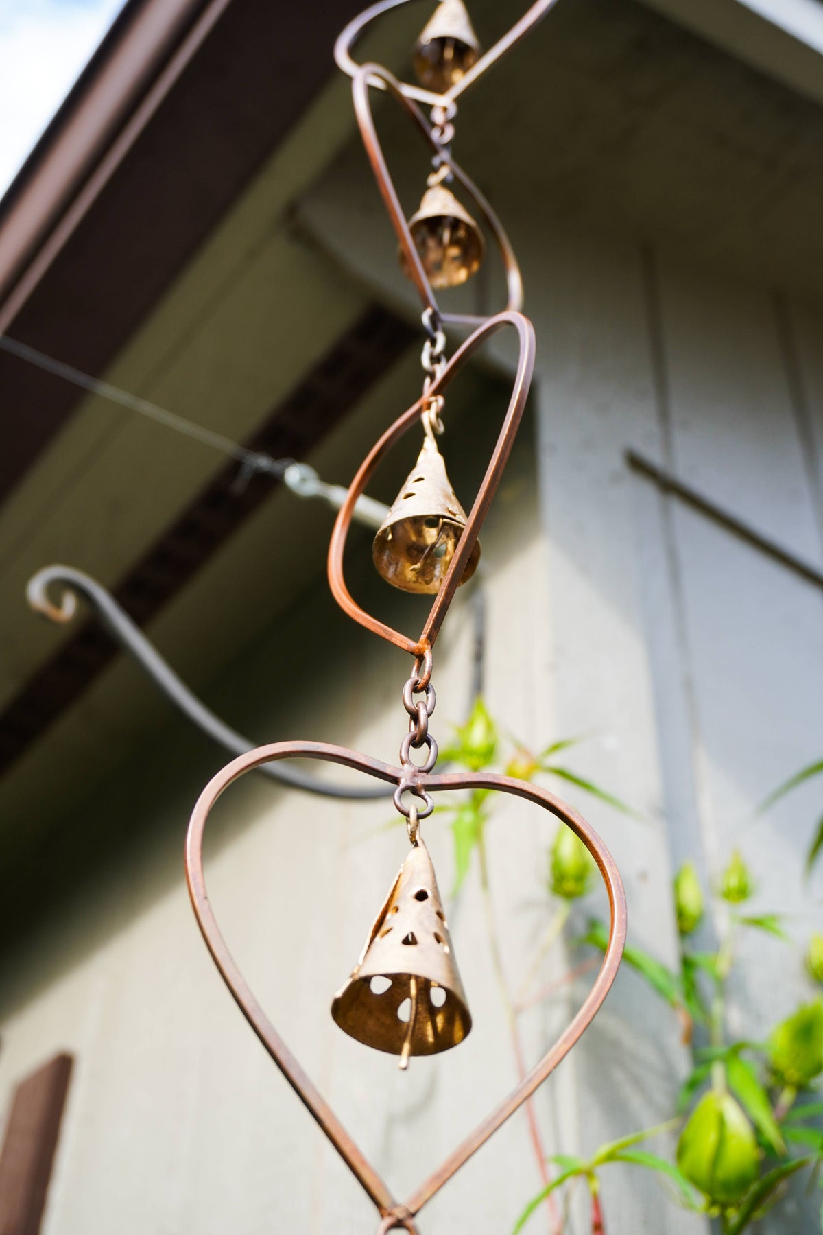 A copper heart-shaped wind spinner with three small hanging bells, photographed outdoors against a house wall and greenery.