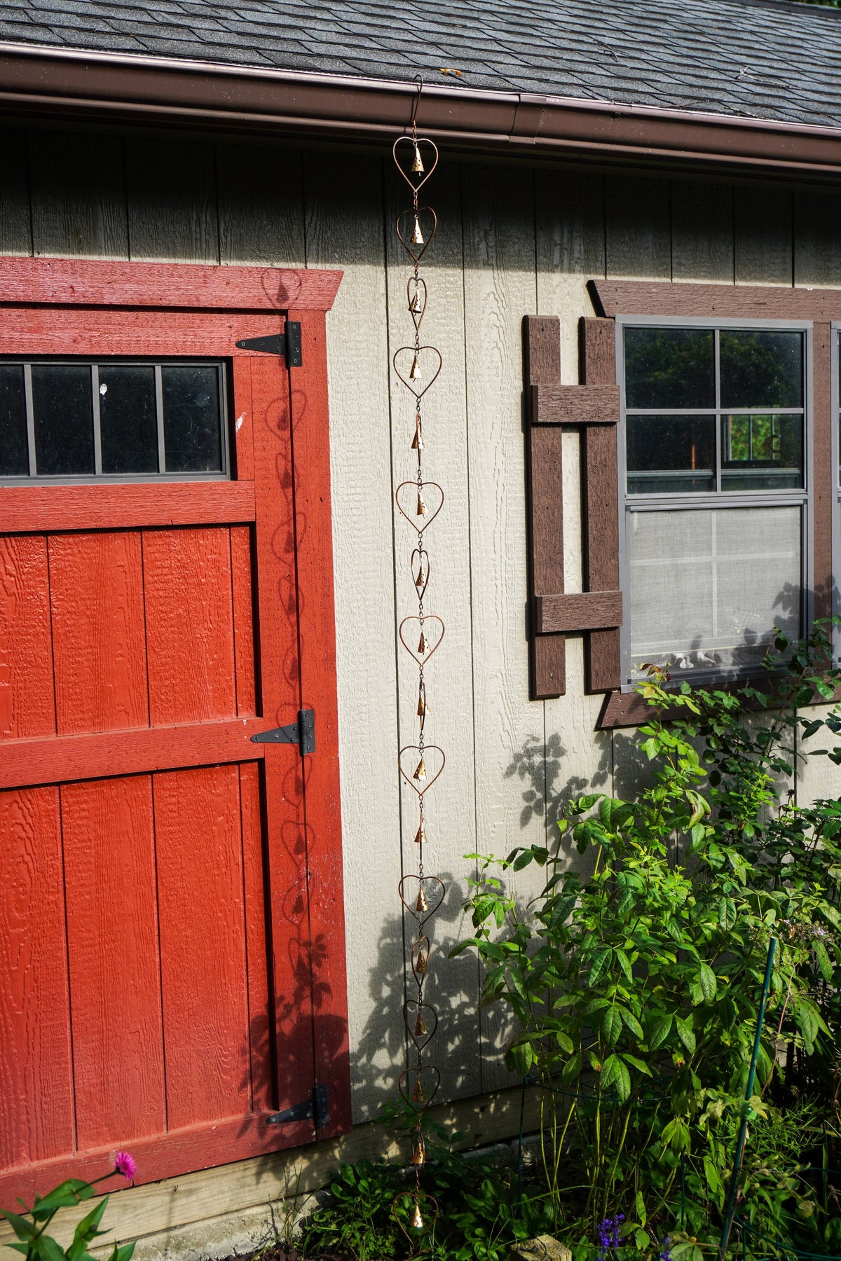 A hanging metal heart-shaped rain chain hangs from the eaves of a house next to a red door and garden plants.