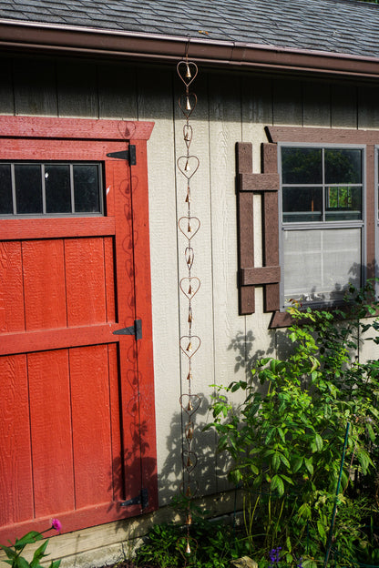 A hanging metal heart-shaped rain chain hangs from the eaves of a house next to a red door and garden plants.