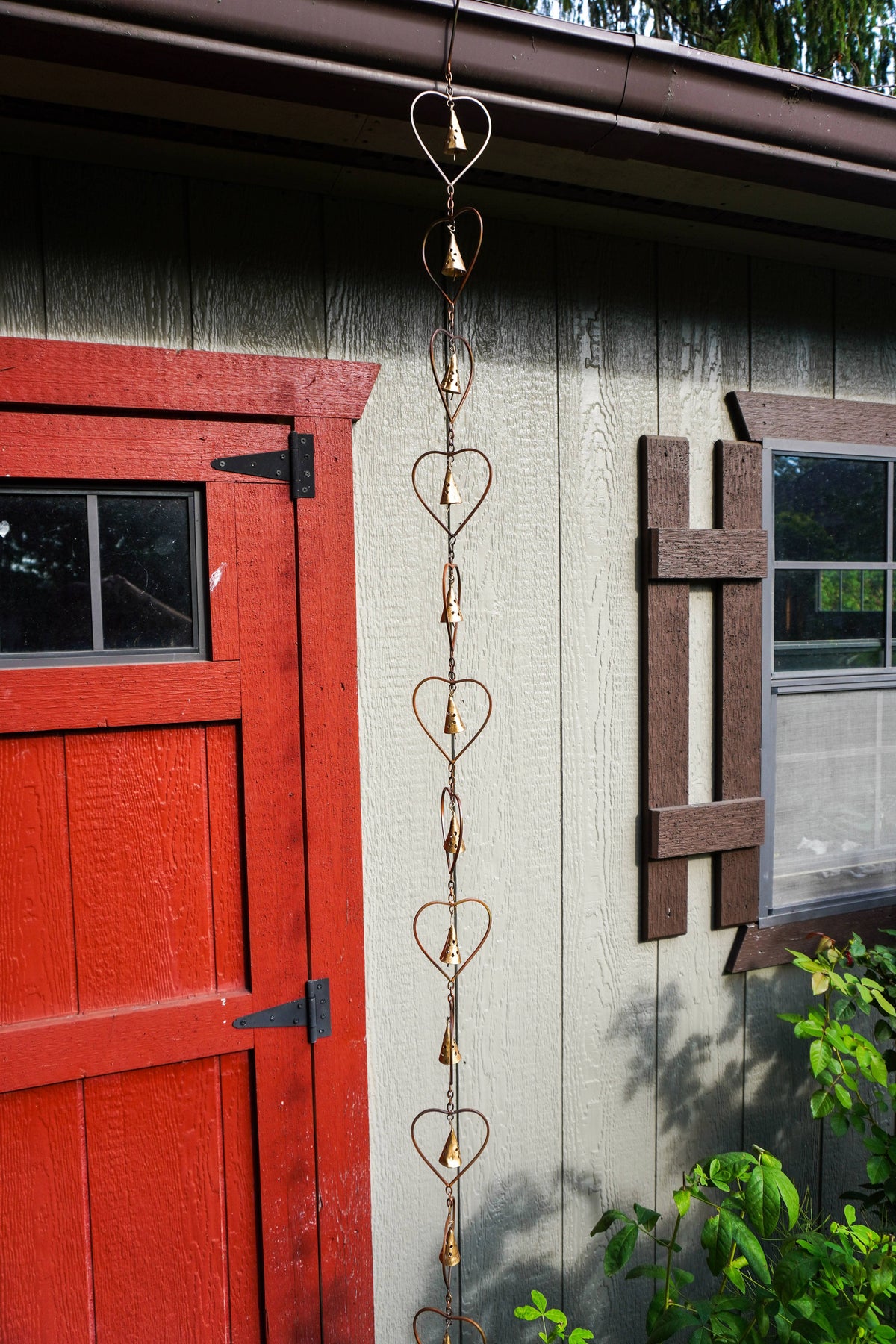 A copper heart-shaped rain chain hangs vertically in front of a house exterior with a red door, beige siding, and a brown window frame.