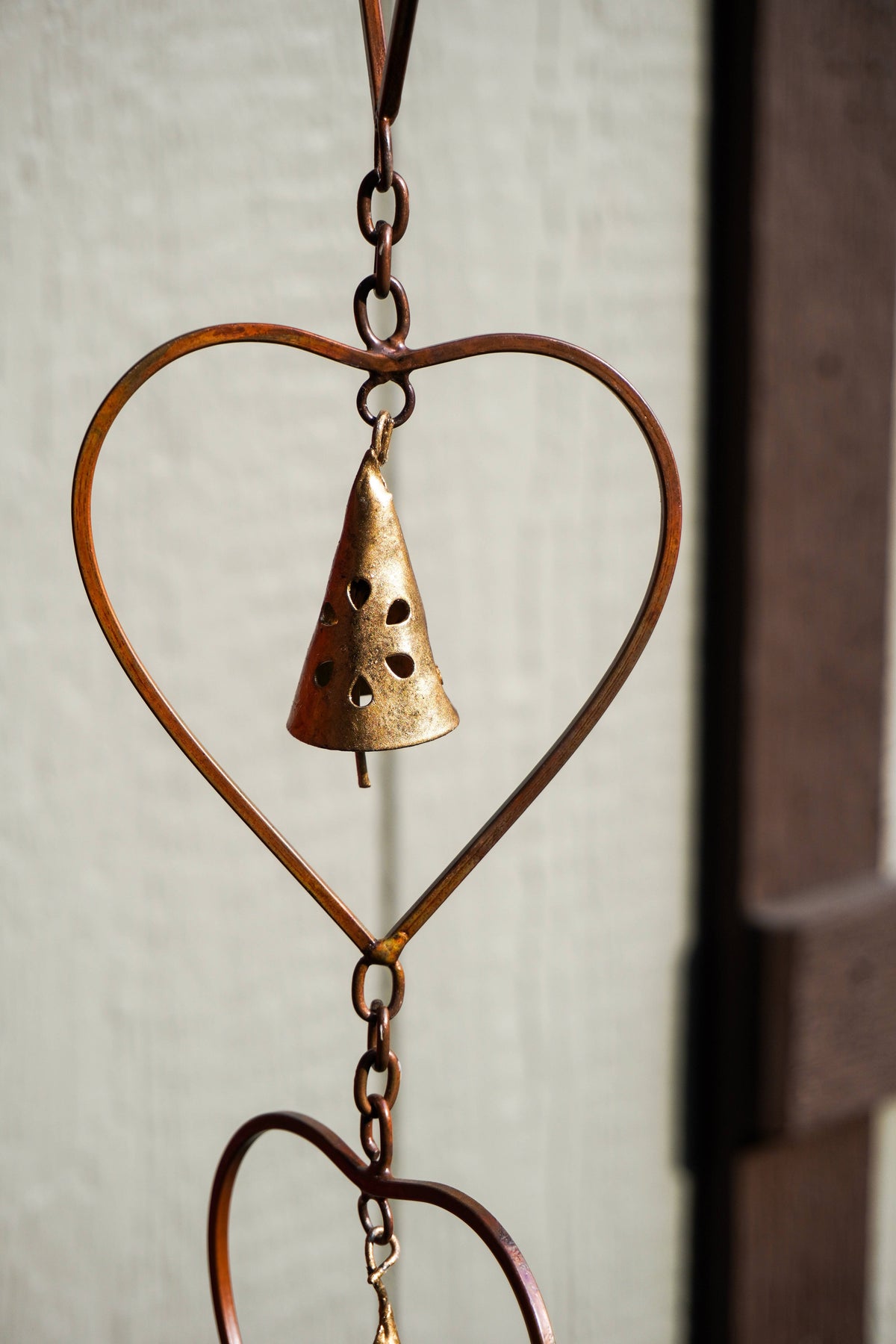 A rustic metal wind chime featuring a large heart-shaped frame and a small hanging bell, photographed against a softly blurred light wall and dark vertical post.