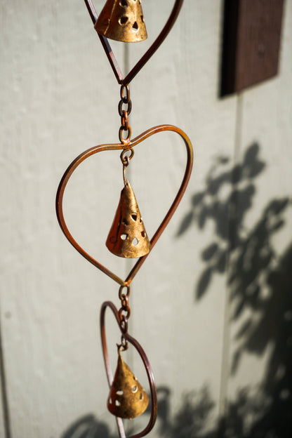 A copper wind spinner featuring a heart-shaped wire frame, perforated cone wind chimes, and a teardrop-shaped ornament, shown hanging against a light wall with leaf shadows.