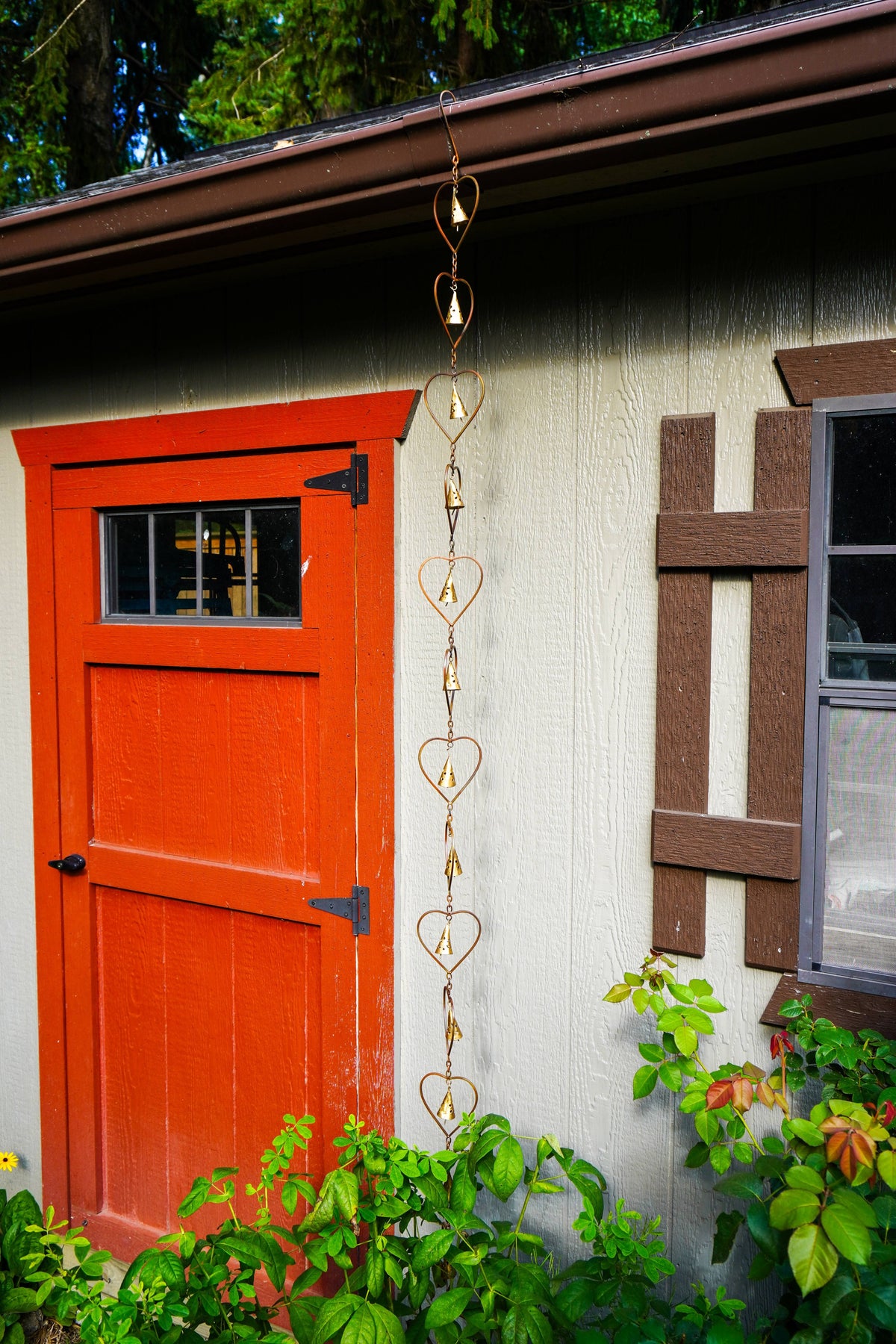 A gold heart-shaped rain chain hangs vertically from the eaves of a house, positioned between a bright orange-red door and a brown window on a beige exterior wall, with green plants at the bottom.