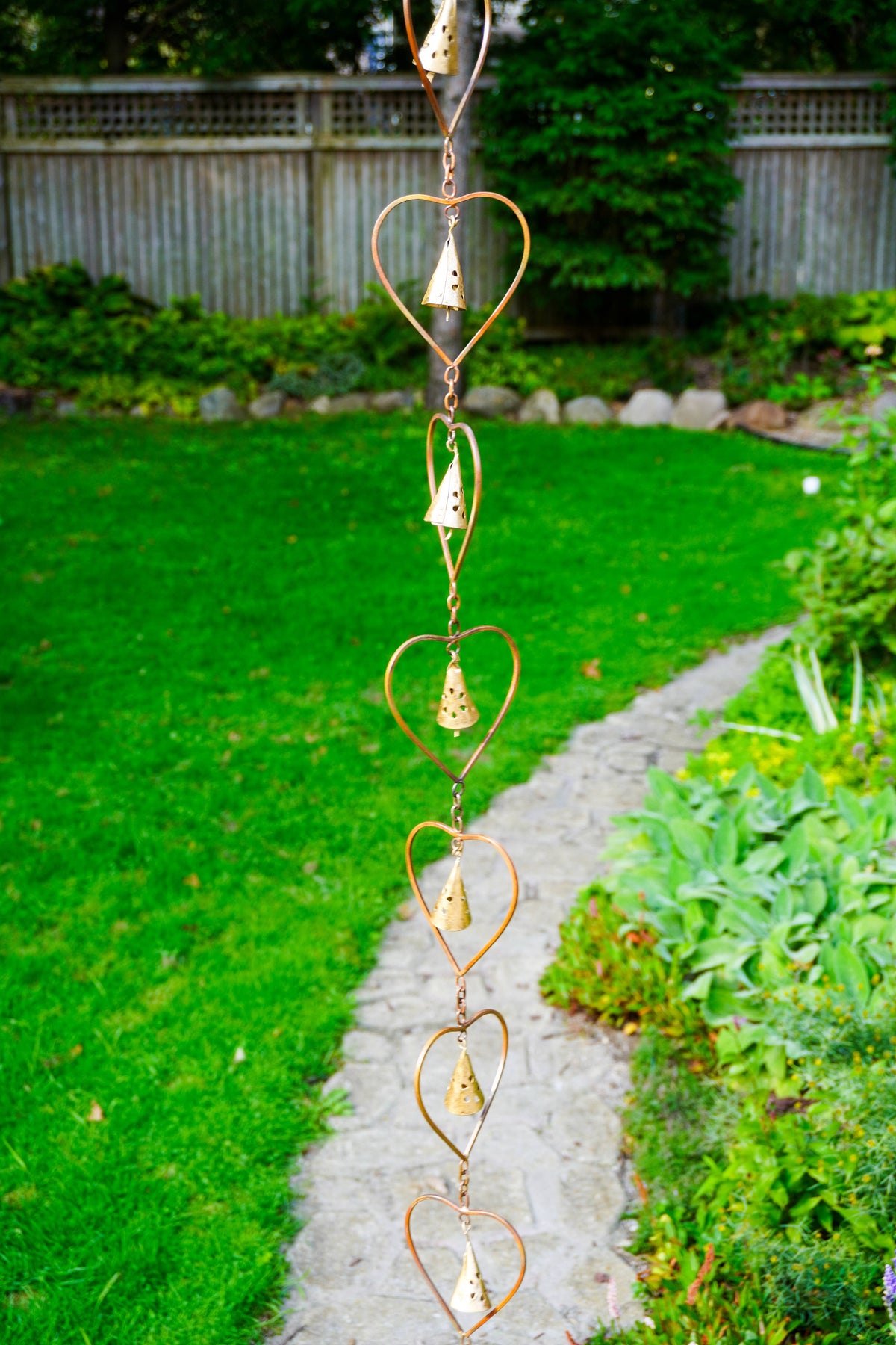 A hanging metal garden wind spinner with a series of heart-shaped wire frames and small bell-like ornaments, photographed outdoors over a garden path with grass and plants in the background.