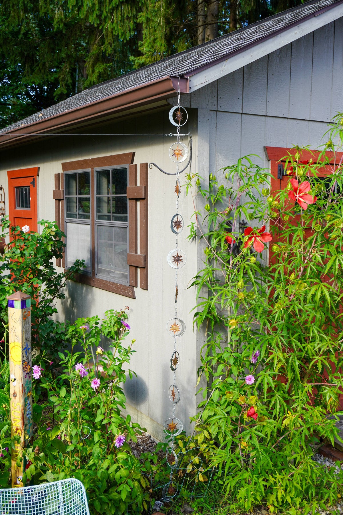 A vertical hanging garden rain chain ornament with multiple circular and star-shaped metal pieces, displayed on the exterior wall of a gray house surrounded by lush green garden plants and flowers.