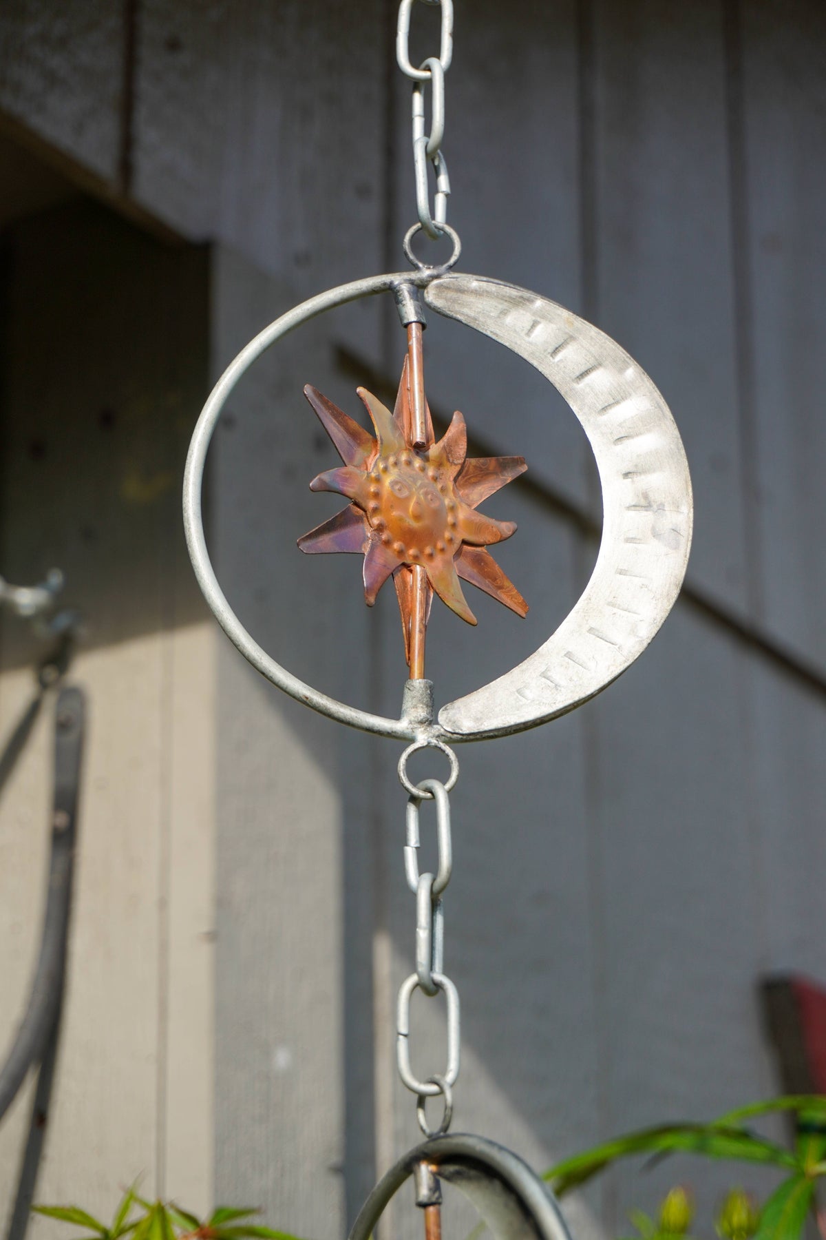 A metal garden wind spinner hanging outdoors against a gray wooden wall. The spinner features a silver crescent moon, a silver outer ring, a bronze sunburst in the center, and hanging chains at the top and bottom.