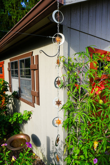 A hanging metal garden star and moon rain chain ornament set displayed outdoors on the exterior wall of a house, surrounded by green plants and flowers.