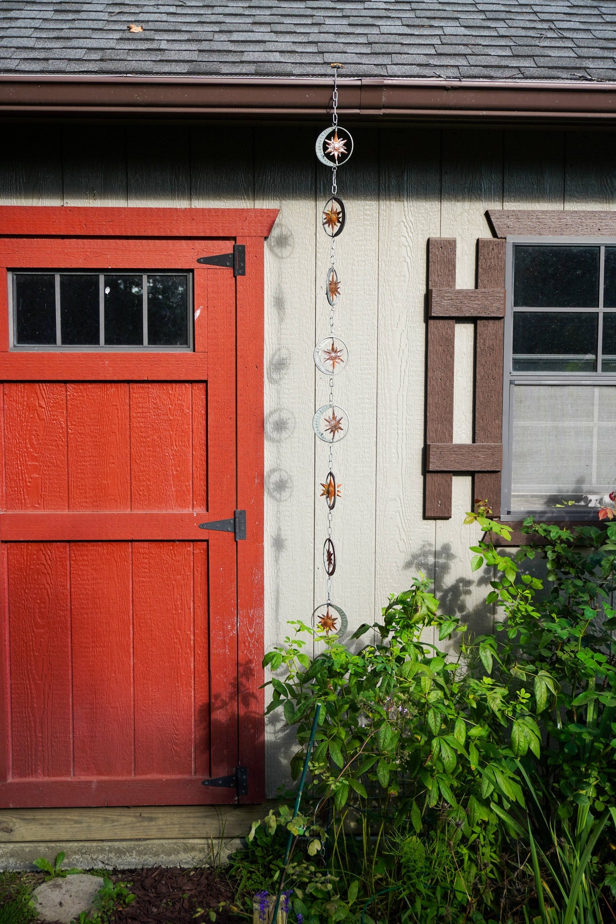 A decorative hanging garden rain chain with sun and moon motifs hangs vertically on the exterior wall of a house next to a red door and green garden plants.
