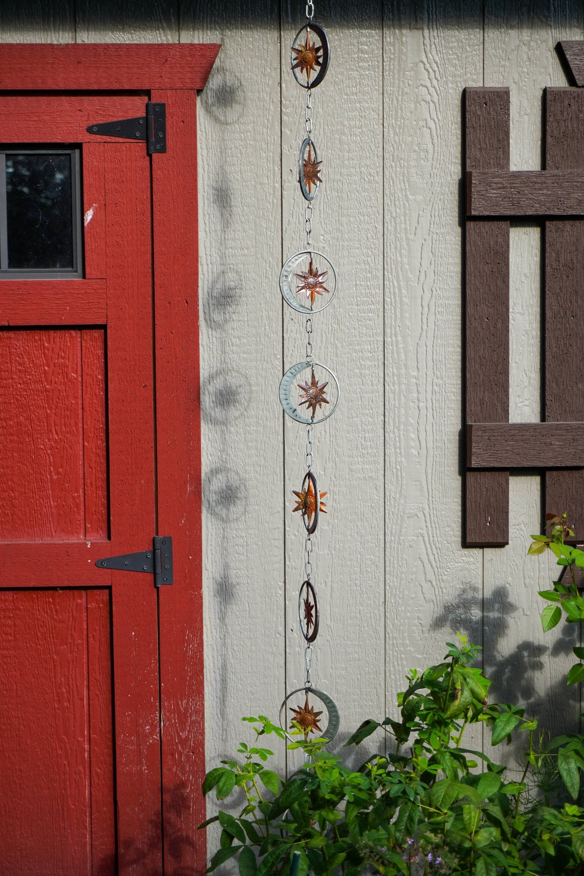 A vertical hanging metal garden rain chain with circular rings and starburst designs, shown outdoors against a beige house wall next to a red door and green plants.