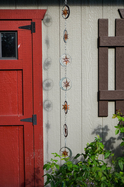 A vertical hanging metal garden rain chain with circular rings and starburst designs, shown outdoors against a beige house wall next to a red door and green plants.