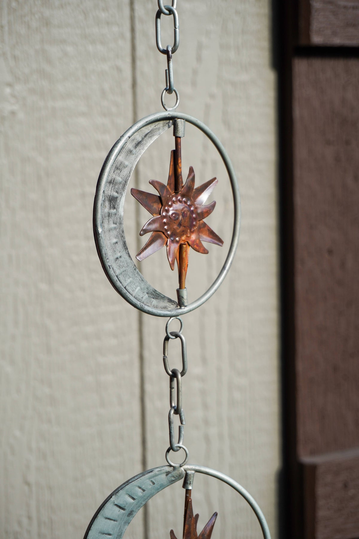 A hanging metal wind spinner with circular rings, a crescent moon, and a starburst design, shown outdoors against a beige wall.