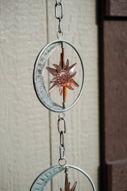 A hanging metal wind spinner with circular rings, a crescent moon, and a starburst design, shown outdoors against a beige wall.