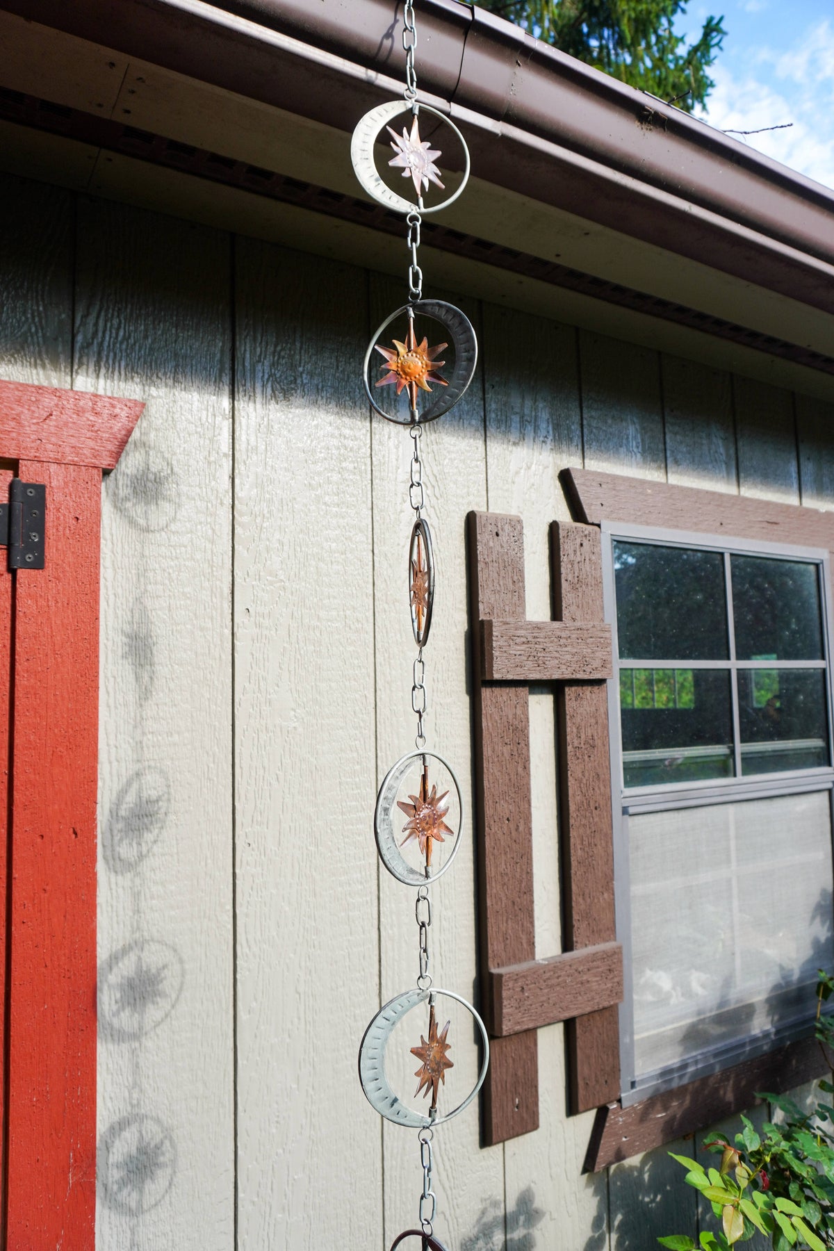 A hanging metal chain ornament with circular rings and starburst compass designs, displayed outdoors against a house exterior wall.