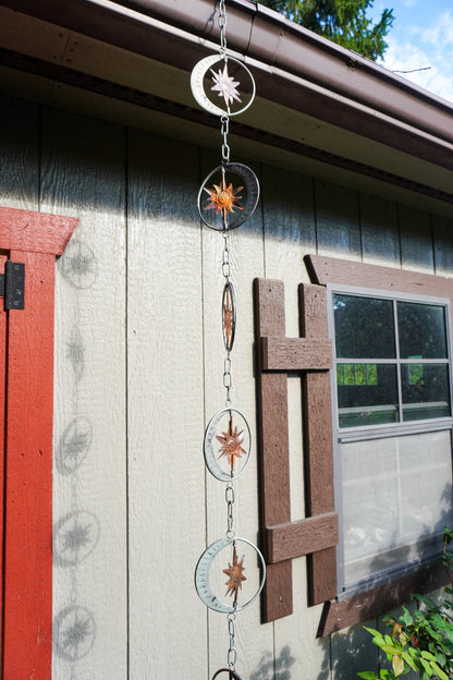 A hanging metal chain ornament with circular rings and starburst compass designs, displayed outdoors against a house exterior wall.